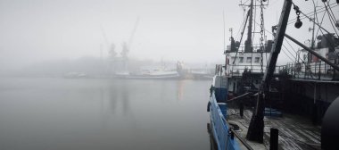 Römorkör ve balıkçı tekneleri (balıkçı tekneleri) limandaki bir rıhtıma demirlemişti. Kalın beyaz sis. Letonya, Baltık Denizi. Panoramik manzara. Hizmet, onarım, nakliye, lojistik, endüstri, ticaret