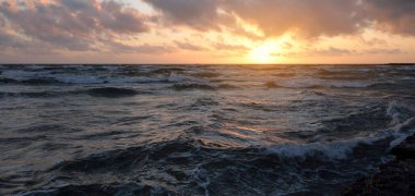 Mavi gökyüzünün altında, parlak pembe ve altın renkli günbatımı bulutlarıyla Baltık deniz kıyısında. Kaya kıyısı, dalgalar. Dalgalar çarpıyor. - Letonya. Doğa, çevre koruma, eko-turizm. Resim gibi bir manzara.