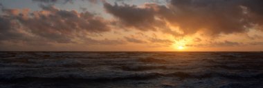 Fırtınadan sonra parlayan pembe ve altın rengi günbatımı bulutlarıyla mavi gökyüzünün altında Baltık Denizi kıyısında. Dalgalar çarpıyor. Doğa, çevre koruma, eko-turizm. Resimli panoramik manzara