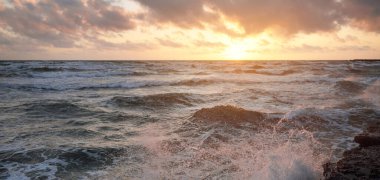 Mavi gökyüzünün altında, parlak pembe ve altın renkli günbatımı bulutlarıyla Baltık deniz kıyısında. Kaya kıyısı, dalgalar. Dalgalar çarpıyor. - Letonya. Doğa, çevre koruma, eko-turizm. Resim gibi bir manzara.
