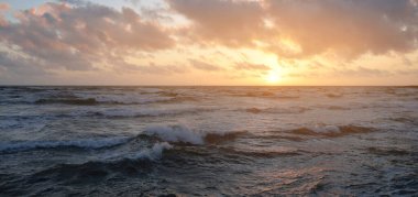 Mavi gökyüzünün altında, parlak pembe ve altın renkli günbatımı bulutlarıyla Baltık deniz kıyısında. Kaya kıyısı, dalgalar. Dalgalar çarpıyor. - Letonya. Doğa, çevre koruma, eko-turizm. Resim gibi bir manzara.