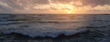 Fırtınadan sonra parlayan pembe ve altın rengi günbatımı bulutlarıyla mavi gökyüzünün altında Baltık Denizi kıyısında. Dalgalar çarpıyor. Doğa, çevre koruma, eko-turizm. Resimli panoramik manzara