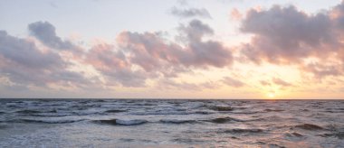 Fırtınadan sonra parlayan günbatımı bulutlarıyla mavi gökyüzünün altında Baltık Denizi kıyısında. Su yüzeyi dokusu, çarpan dalgalar. Doğa, çevre koruma, eko-turizm. Resimli panoramik manzara