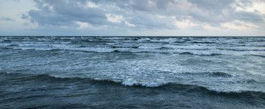 Fırtınadan sonra parlayan günbatımı bulutlarıyla mavi gökyüzünün altında Baltık Denizi kıyısında. Su yüzeyi dokusu, çarpan dalgalar. Doğa, çevre koruma, eko-turizm. Resimli panoramik manzara