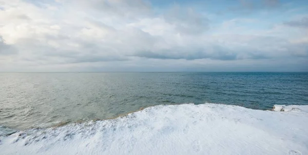 Panoramic view from the snow-covered Baltic sea shore at sunset. Latvia. Dramatic cloudscape. Fickle weather, waves and water splashes. Winter tourism, global warming, climate change, nature, ecology