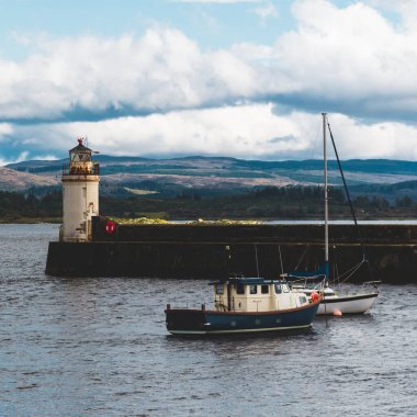 Mavi balıkçı teknesi ve bulutlu bir günde yelken açan bir yat, yakın plan. Arka planda kayalık kıyılar var. Dramatik bulut manzarası. Ardrishaig, Crinan Kanalı, İskoçya, İngiltere. Seyahat yerleri, gemi yolculuğu.