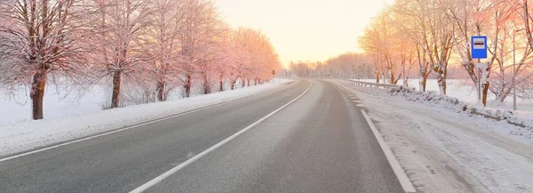 Karla kaplı orman ve kırsal alanda boş otoyol (asfalt yol). Otobüs durağı. Gündoğumu, kar yağışı. Doğa, Noel tatilleri, uzak yerler, kış lastikleri, tehlikeli sürüş konsepti