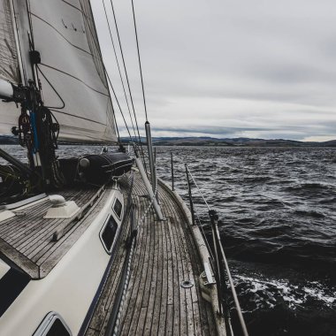 Bulutlu bir günde Tarbert 'in kayalık kıyılarında seyir halindeki ahşap meşe güvertesiyle donatılmış modern bir yat. İskoçya, İngiltere. Güverteden pruvaya, direğe ve yelkenlere yakın görüntü. Seyahat yerleri