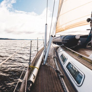 Yağmurdan sonra Tarbert 'in kayalık kıyılarında seyir halindeki ahşap meşe güvertesiyle donatılmış modern bir yat. İskoçya, İngiltere. Güverteden pruvaya, direğe ve yelkenlere yakın görüntü. Açık mavi gökyüzü