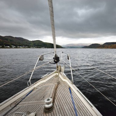 Bulutlu bir günde ahşap meşe güverteli, yelkenli, modern bir yat. Güverteden pruvaya bir manzara. Bute 'un kayalık kıyılarının sudan panoramik görüntüsü. Tepeler ve dağlar arka planda. Bute Adası, Clyde Firth, İskoçya