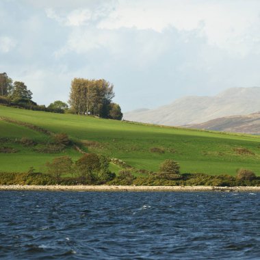 Sudan kayalık nehir kıyılarının panoramik görüntüsü. Arka planda ağaçlar, tepeler ve dağlar var. Bulutlu mavi gökyüzü. Gare Loch, Clyde Firth, İskoçya, İngiltere