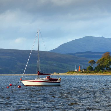 Rhu marinasında demirli yelkenli bir yat. Arkasında tepeler, ormanlar ve kır evleri olan kayalık kıyılar. Clyde Firth, İskoçya, İngiltere