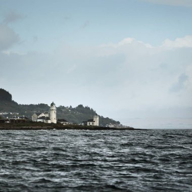 Sudan kayalık nehir kıyılarının panoramik görüntüsü. Arka planda ağaçlar, tepeler ve dağlar var. Deniz feneri ve kır evleri. Bulutlu mavi gökyüzü. Gare Loch, Clyde Firth, İskoçya, İngiltere