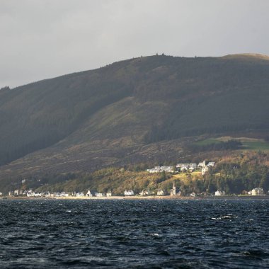 Sudan kayalık nehir kıyılarının panoramik görüntüsü. Arka planda kır evleri, ağaçlar, tepeler ve dağlar var. Bulutlu mavi gökyüzü. Gare Loch, Clyde Firth, İskoçya, İngiltere
