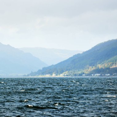 Sudan kayalık nehir kıyılarının panoramik görüntüsü. Arka planda kır evleri, ağaçlar, tepeler ve dağlar var. Bulutlu mavi gökyüzü. Gare Loch, Clyde Firth, İskoçya, İngiltere