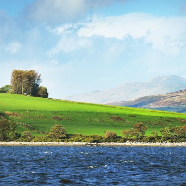 Sudan kayalık nehir kıyılarının panoramik görüntüsü. Arka planda ağaçlar, tepeler ve dağlar var. Bulutlu mavi gökyüzü. Gare Loch, Clyde Firth, İskoçya, İngiltere