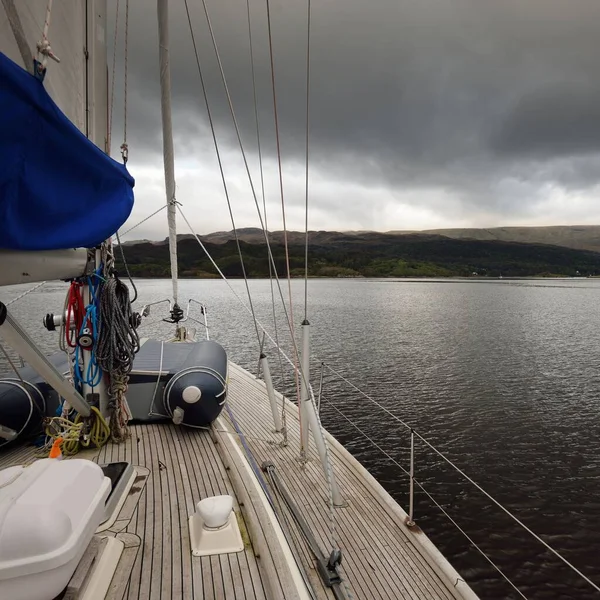 Sloop rigged modern yacht with wooden teak deck sailing on a cloudy day. A view from the deck to the bow. Panoramic view of the rocky shores of Kyles of Bute from the water. Hills and mountains in the background. Bute island, Firth of Clyde, Scotland
