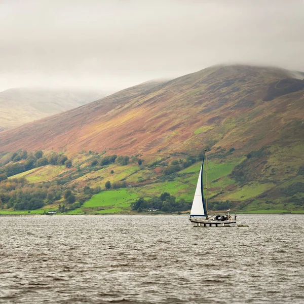 Sloop rigged yacht with a motor boat sailing on a cloudy day. Panoramic view of the rocky shores of Kyles of Bute from the water. Hills and mountains in the background. Bute island, Firth of Clyde, Scotland, UK