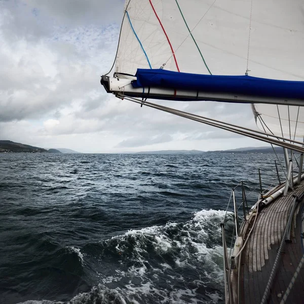 Sloop rigged modern yacht with wooden teak deck sailing  in the Firth of Clyde. Forests, hills and mountains of the Bute Island in the background. Dark stormy sky. Scotland, UK