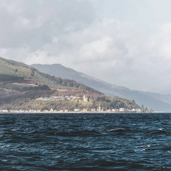 Village on the rocky shore of the Firth of Clyde river, panoramic view from a sailing boat. Dramatic sky. Scotland, UK. Travel destinations, eco tourism, vacations, pure nature. Atmospheric landscape