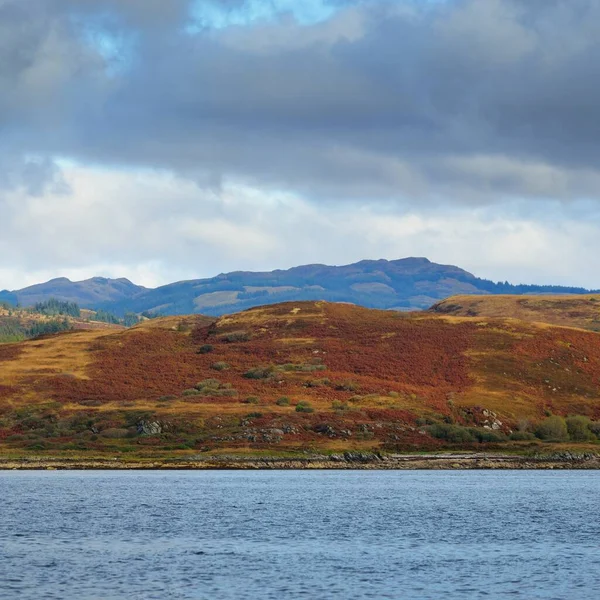 Sudan kayalık nehir kıyılarının panoramik görüntüsü. Arka planda ağaçlar, tepeler ve dağlar var. Bulutlu mavi gökyüzü. Gare Loch, Clyde Firth, İskoçya, İngiltere