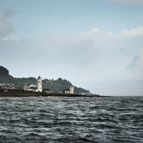 Sudan kayalık nehir kıyılarının panoramik görüntüsü. Arka planda ağaçlar, tepeler ve dağlar var. Deniz feneri ve kır evleri. Bulutlu mavi gökyüzü. Gare Loch, Clyde Firth, İskoçya, İngiltere