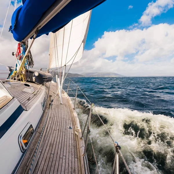 Sloop rigged modern yacht with wooden teak deck sailing near the rocky shore of the Firth of Clyde river. Dramatic sky. Scotland, UK. Travel destinations, eco tourism, vacations, pure nature