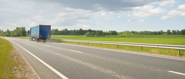 Modern semi-trailer truck riding through green plowed agricultural field and forest. Highway, freight transportation, articulated lorry, logistics, industry, delivery, remote places, route. Europe