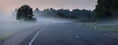 Gün batımında sisli bir yolda (asfalt yol). Ayın doğuşu, alacakaranlık, dramatik gökyüzü. Idyllic yaz kırsal manzarası. Ulaşım, uzak yerler, tehlikeli sürüş