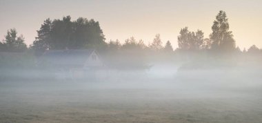 Gün batımında bir siste kır evi ve tarlaları (orman çimleri, çayır). Ayın doğuşu, alacakaranlık, açık gökyüzü. Idyllic yaz kırsal manzarası. Doğa, çevre, eko-turizm, uzak yerler, tatiller