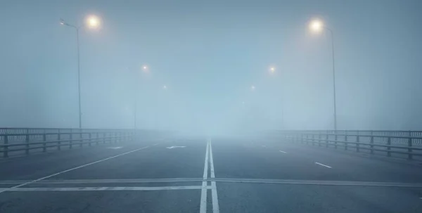 An empty illuminated asphalt road (highway) in a thick fog. Pedestrian walkway, crossing, street lights. Dangerous driving, walking, cycling, traffic laws concepts
