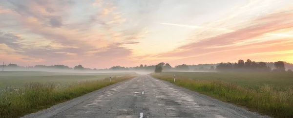 Country highway (old asphalt road) through the field and forest at sunrise. Soft sunlight, glowing pink and golden clouds. Vacations, logistics, remote places