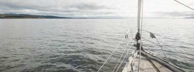 Kayalık göl kıyılarında seyir halindeki ahşap meşe güvertesiyle donatılmış modern bir yat. İskoçya, İngiltere. Güverteden pruvaya, direğe, yelkenlere bakın. Seyahat tutkusu, eğlence aktivitesi, spor, yelkenli yarışı, deniz yolculuğu.