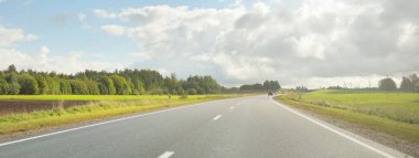 Country highway (asphalt road) through the fields and forest. View from the car. Dramatic storm clouds. Travel destinations, route, vacations, logistics, remote places