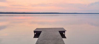 Forest lake at sunset. Wooden pier. Soft sunlight, glowing clouds, symmetry reflections in a crystal clear water. Idyllic landscape. Panoramic view. Nature, ecology, ecotourism. Peace and joy concepts