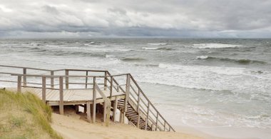 Fırtınadan sonra Baltık kıyısı (plaj). Modern tahta kaldırım (tahta kaldırım). Kum tepeleri. Dramatik gökyüzü, dalgalar ve su sıçramaları. Doğa, ekoloji, eko-turizm, yürüyüş, eğlence kavramları