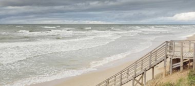 Fırtınadan sonra Baltık kıyısı (plaj). Modern tahta kaldırım (tahta kaldırım). Kum tepeleri. Dramatik gökyüzü, dalgalar ve su sıçramaları. Doğa, ekoloji, eko-turizm, yürüyüş, eğlence kavramları