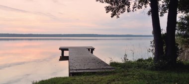 Forest lake at sunset. Wooden pier. Soft sunlight, glowing clouds, symmetry reflections in a crystal clear water. Idyllic landscape. Panoramic view. Nature, ecology, ecotourism. Peace and joy concepts