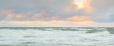 Gün batımında Baltık Denizi. Dramatik gökyüzü, mavi ve pembe parlayan bulutlar, altın güneş ışığı. Dalgalar, su sıçratma. Resimli manzara, deniz manzarası, doğa. Panoramik görünüm, uzun pozlama