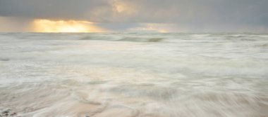 Gün batımında Baltık Denizi. Dramatik gökyüzü, mavi ve pembe parlayan bulutlar, altın güneş ışığı. Dalgalar, su sıçratma. Resimli manzara, deniz manzarası, doğa. Panoramik görünüm, uzun pozlama