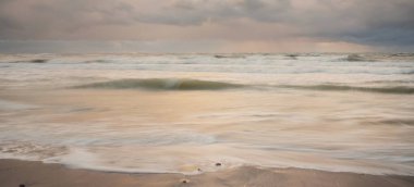 Gün batımında Baltık Denizi. Dramatik gökyüzü, mavi ve pembe parlayan bulutlar, altın güneş ışığı. Dalgalar, su sıçratma. Resimli manzara, deniz manzarası, doğa. Panoramik görünüm, uzun pozlama