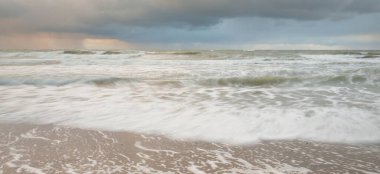 Gün batımında Baltık Denizi. Dramatik gökyüzü, mavi ve pembe parlayan bulutlar, altın güneş ışığı. Dalgalar, su sıçratma. Resimli manzara, deniz manzarası, doğa. Panoramik görünüm, uzun pozlama