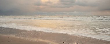 Gün batımında Baltık Denizi. Dramatik gökyüzü, mavi ve pembe parlayan bulutlar, altın güneş ışığı. Dalgalar, su sıçratma. Resimli manzara, deniz manzarası, doğa. Panoramik görünüm, uzun pozlama