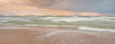 Gün batımında Baltık Denizi. Dramatik gökyüzü, mavi ve pembe parlayan bulutlar, altın güneş ışığı. Dalgalar, su sıçratma. Resimli manzara, deniz manzarası, doğa. Panoramik görünüm, uzun pozlama