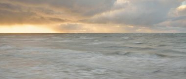 Gün batımında Baltık Denizi. Dramatik gökyüzü, mavi ve pembe parlayan bulutlar, altın güneş ışığı. Dalgalar, su sıçratma. Resimli manzara, deniz manzarası, doğa. Panoramik görünüm, uzun pozlama