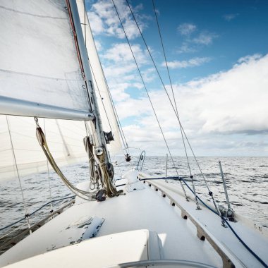 Açık bir günde açık denizde yelken açan beyaz yelkenli bir yat. Güverteden pruvaya bir manzara. Kümülüs bulutları. Ulaşım, seyahat, gezi, spor, eğlence, boş zaman aktivitesi, yarış, yelkenli yarışı
