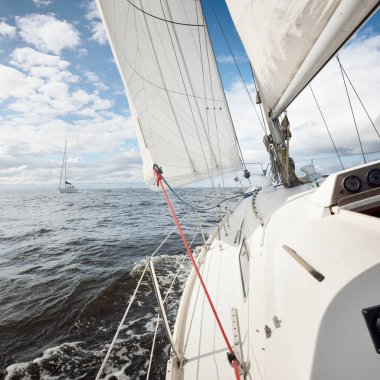 Açık bir günde açık denizde yelken açan beyaz yelkenli bir yat. Güverteden pruvaya bir manzara. Kümülüs bulutları. Ulaşım, seyahat, gezi, spor, eğlence, boş zaman aktivitesi, yarış, yelkenli yarışı