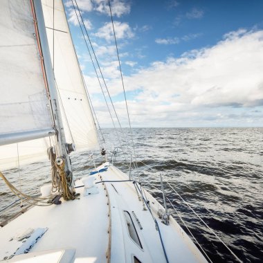 Açık bir günde açık denizde yelken açan beyaz yelkenli bir yat. Güverteden pruvaya bir manzara. Kümülüs bulutları. Ulaşım, seyahat, gezi, spor, eğlence, boş zaman aktivitesi, yarış, yelkenli yarışı
