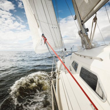 Açık bir günde açık denizde yelken açan beyaz yelkenli bir yat. Güverteden pruvaya bir manzara. Kümülüs bulutları. Ulaşım, seyahat, gezi, spor, eğlence, boş zaman aktivitesi, yarış, yelkenli yarışı