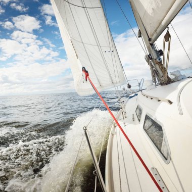 Açık bir günde açık denizde yelken açan beyaz yelkenli bir yat. Güverteden pruvaya bir manzara. Kümülüs bulutları. Ulaşım, seyahat, gezi, spor, eğlence, boş zaman aktivitesi, yarış, yelkenli yarışı